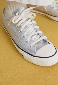 Zapatillas de lona plateadas metálicas con punteras de goma blanca, cordones planos y acentos negros en los laterales sobre una superficie amarilla texturizada.