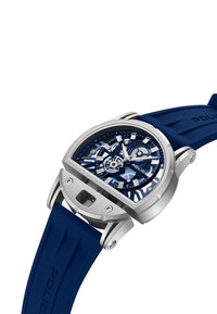 Ura z modrim silikonskim paščkom s srebrnim ohišjem iz nerjavečega jekla, okvirom z osvetljenim mehanizmom in zapletenimi modrimi detajli. Prikazuje športen, moderen dizajn.