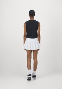Top negro sin mangas combinado con una falda corta de vuelo blanca. La modelo lleva zapatillas deportivas negras y calcetines blancos hasta el tobillo, vista desde atrás.