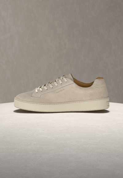 Μπεζ καστόρινο sneaker χαμηλού ύψους με λευκά κορδόνια και χοντρή λαστιχένια σόλα, απεικονίζεται σε φωτεινή υφή επιφάνειας σε ουδέτερο φόντο.