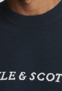 Maglione lavorato a maglia blu navy con scollo rotondo, dotato della scritta bianca "LITTLE & SCOTT" in evidenza sul petto. Texture liscia.