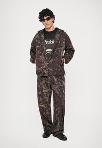 Giacca e pantaloni camouflage in tonalità terrose di marrone e bordeaux, abbinati a una maglietta nera con grafica e occhiali da sole scuri.