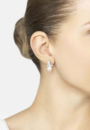 Woman wearing silver butterfly-shaped earrings with clear stones and a hanging round white pearl, hair tied back against a white background.