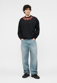 Jeune homme aux cheveux bouclés portant un sweat-shirt noir avec un texte rouge, un jean bleu clair ample et des baskets noires, debout les mains dans les poches.
