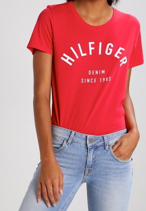 Osoba nosząca czerwoną koszulkę z krótkim rękawem z białym napisem ""HILFIGER DENIM SINCE 1985"" oraz jasnoniebieskie dżinsy z jedną ręką w kieszeni.