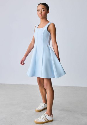 Fiatal személy, aki éppen áll, világoskék ujjatlan, bővülő szoknyás ruhát és fehér tornacipőt visel bézs talppal, szürke padlón.