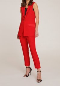 Blazer sans manches rouge avec poches avant, assorti à un pantalon rouge ajusté. Talons hauts noirs à lanières avec brides au niveau de la cheville, en tissu lisse.