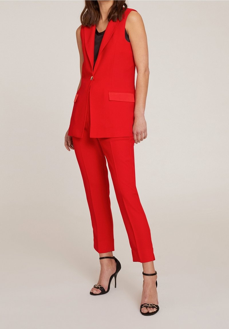 Blazer sans manches rouge avec poches avant, assorti à un pantalon rouge ajusté. Talons hauts noirs à lanières avec brides au niveau de la cheville, en tissu lisse.