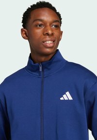 Jūras spēku zip-up jaka, izgatavota no mīksta auduma ar augstu apkakli un balto Adidas logo kreisajā krūtī. Arī izcila šuvuma detaļas un pieguļošs dizains.