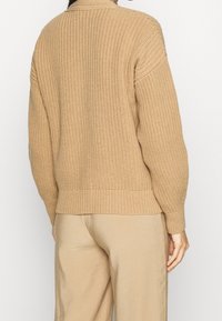 Beige neulepusero, jossa on teksturoitu kuvio, pitkät hihat ja ribbireunus. Takana näkyy rento istuvuus ja korkea kaulus.