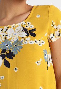 Bluse mit Rundhalsausschnitt in Gelb mit blau-weißer Blumenmusterung; leichtes, strukturiertes Gewebe; kurze Ärmel; Blumenakzente entlang des Ausschnitts.