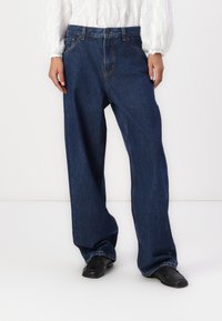 Bredde ben blå denimjeans med en falmet finish. Har en knappelukking og fem-lommers design. Stylet med en hvit bluse og svarte sko.
