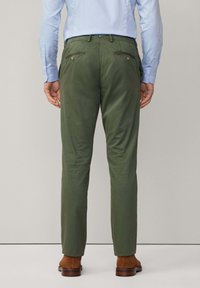 Zelene hlače od tkanine, slim fit kroja, s stražnjim džepovima s kopčama i blagim sjajem. Uparene s smeškim obućom.
