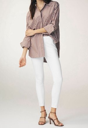 Femme portant une chemise oversize rayée marron et blanc avec manches retroussées, pantalon slim blanc et sandales à talons marron, debout.