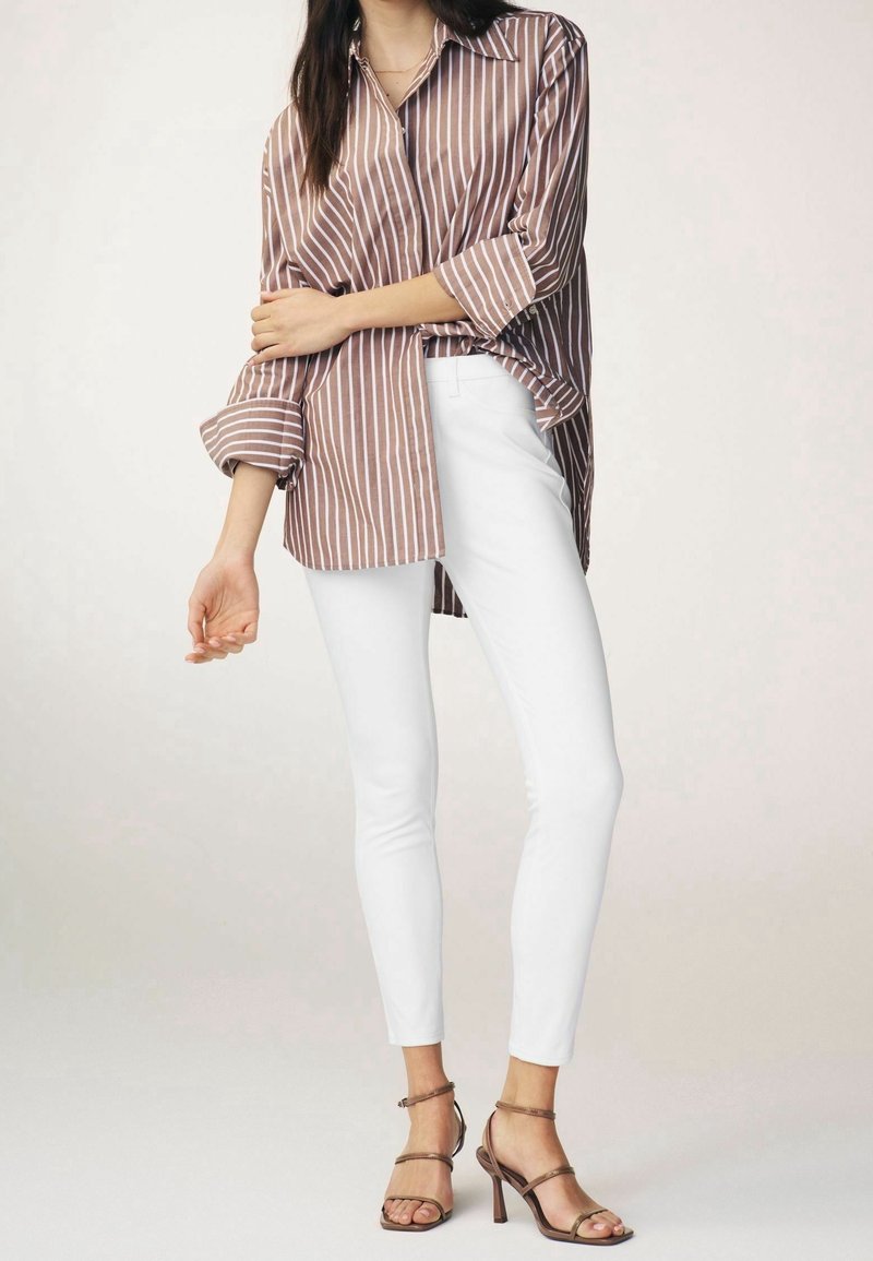 Femme portant une chemise oversize rayée marron et blanc avec manches retroussées, pantalon slim blanc et sandales à talons marron, debout.