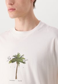 Balta medvilninė marškinėliai su žaliu palmės medžio grafikos piešiniu ir juodu tekstu "La La La" bei "West Coast Stories Since 1993".