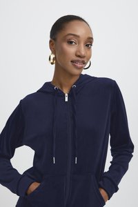 Marinblå zip-up hoodie gjord av mjukt sammetsmaterial, med dragsko i huvan, sidofickor och silverfärgade detaljer.