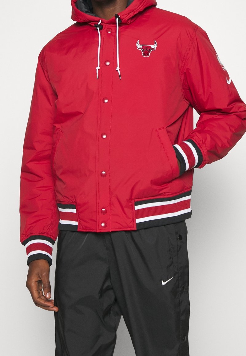 Homme portant une veste rouge Chicago Bulls avec des poignets rayés noirs, blancs et rouges et un pantalon Nike noir avec logo blanc.