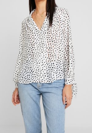 Witte blouse met lange mouwen en knopen met zwarte stippen, met gestrikte manchetten, gecombineerd met lichtblauwe spijkerbroek en gelaagde gouden kettingen.