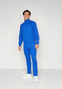 Μπλε φόρμα από ελαστικό υλικό με λευκές ρίγες στα πλάγια, που περιλαμβάνει ζακέτα με φερμουάρ και ταιριαστό παντελόνι. Διακοσμημένη με ένα λογότυπο.