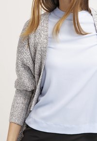 Top azzurro chiaro a maniche corte con finitura testurizzata, indossato sotto un morbido cardigan grigio a maglia aperta. Entrambi i capi hanno vestibilità comoda.