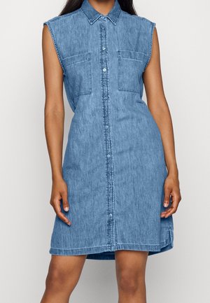 Ærmeløs blå denimkjole med knapper foran, krave og to brystlommer, båret af en person stående mod en ensfarvet baggrund.