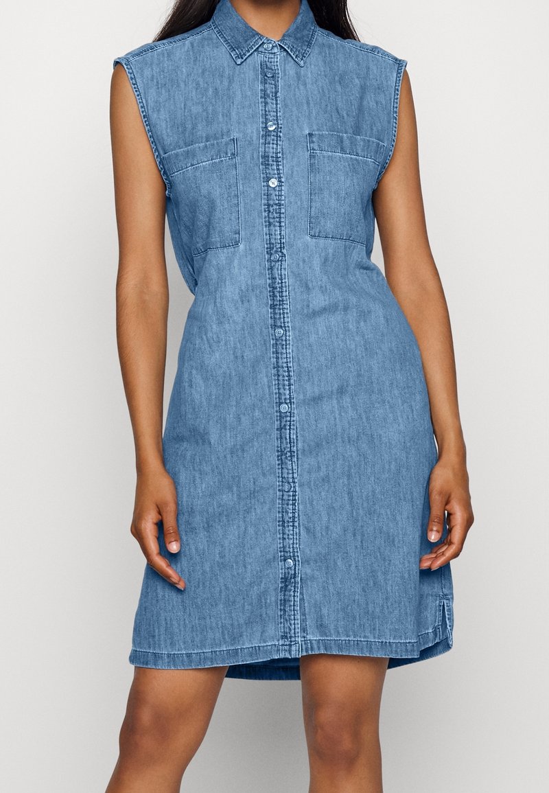Robe en jean bleue sans manches avec patte de boutonnage à l’avant, col et deux poches poitrine portée par une personne debout devant un fond uni.