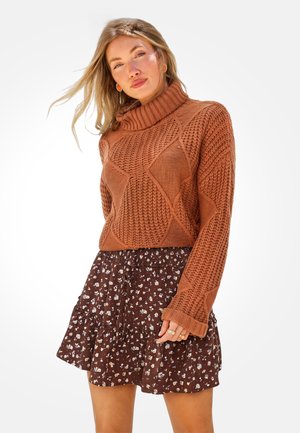 Woman with long blonde hair wearing a rust-colored chunky knit turtleneck sweater and a brown floral ruffled skirt.