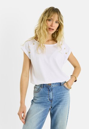 Mujer rubia con cabello ondulado, que lleva una camisa blanca de manga corta con botones y unos vaqueros de tiro alto en azul claro, posando con la mano en el bolsillo.