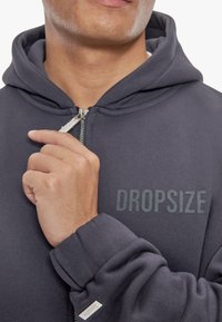 Mørk grå zip-up hættetrøje med en blød tekstur, der har et trykt "DROPSIZE" logo. Sølvfarvet lynlås hardware og ribbede manchetter er bemærkelsesværdige.