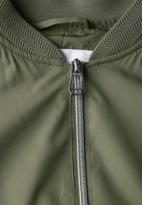 Giacca bomber verde oliva con colletto a coste, dotata di una zip argentata e una texture del tessuto liscia. Inquadratura ravvicinata della zip e del dettaglio del colletto.