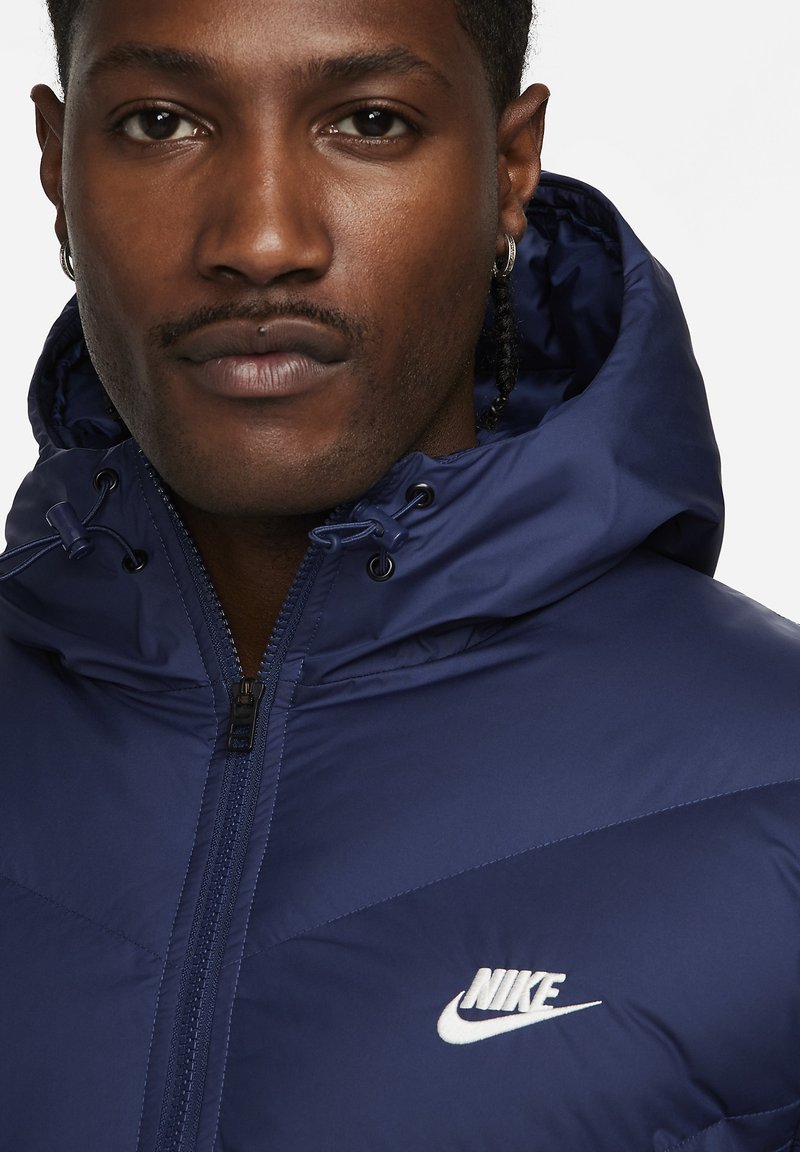 Marineblaue isolierte Jacke mit Kapuze, ausgestattet mit einem Reißverschluss auf der Vorderseite und dem weißen gestickten Nike-Logo auf der Brust. Glattes, wasserabweisendes Material.