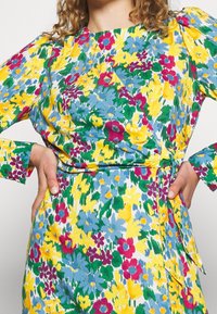 Blommig omlottklänning med ett livligt mönster av gula, gröna, rosa och blå blommor, långa ärmar och en knytning i midjan.