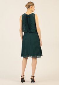 Faltendes grünes Kleid mit einem geschichteten Design und transparentem Rücken, versehen mit einem runden Ausschnitt und einer Länge über dem Knie. Akzentuiert mit Absatzsandalen.