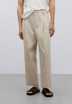 Personne portant un pantalon large beige et des sandales marron foncé debout sur un sol gris devant un mur uni gris clair.