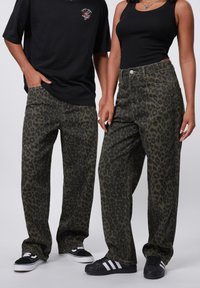 Leopard print jeans in dark green and black, featuring a loose fit, five-pocket design, and metal button accent. Black sneakers worn.