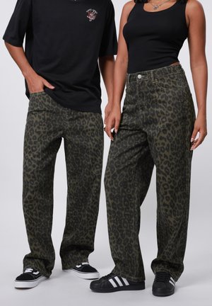 Leopardskindsjeans i mørkegrøn og sort, med en løs pasform, fem-lomme design og metalknap detalje. Sorte sneakers båret.