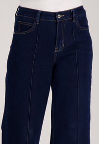 Donkerblauwe spijkerbroek van denim met een hoge taille, voorzakken en contrasterende gouden stiksels langs de naden.