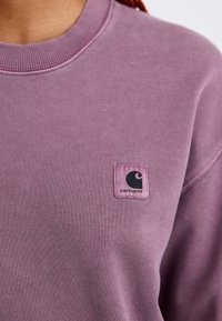 Close-up of a person wearing a dusty purple Carhartt sweatshirt with a small black logo patch on the chest.