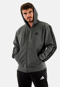 Felpa grigia con zip, cappuccio regolabile con cordino, dettagli neri a righe sulle maniche e tasche frontali. Presenta un logo Adidas sul petto.