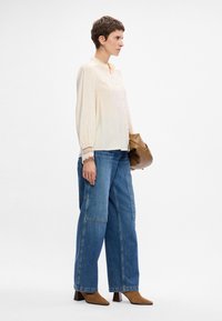 Blusa crema con chiusura a bottoni e maniche rifinite in pizzo, abbinata a jeans blu larghi e stivali con tacco color cuoio. Borsa a mano marrone come accessorio.