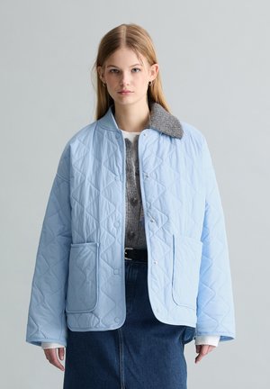Jeune femme portant une veste matelassée bleu clair avec col gris, un pull gris et une jupe en jean foncé, debout devant un fond uni.