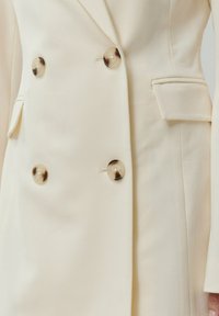 Doppelt breiter cremefarbener Blazer mit großen goldfarbenen Knöpfen, ausgestattet mit einer einzelnen Brusttasche und einer glatten Stofftextur.