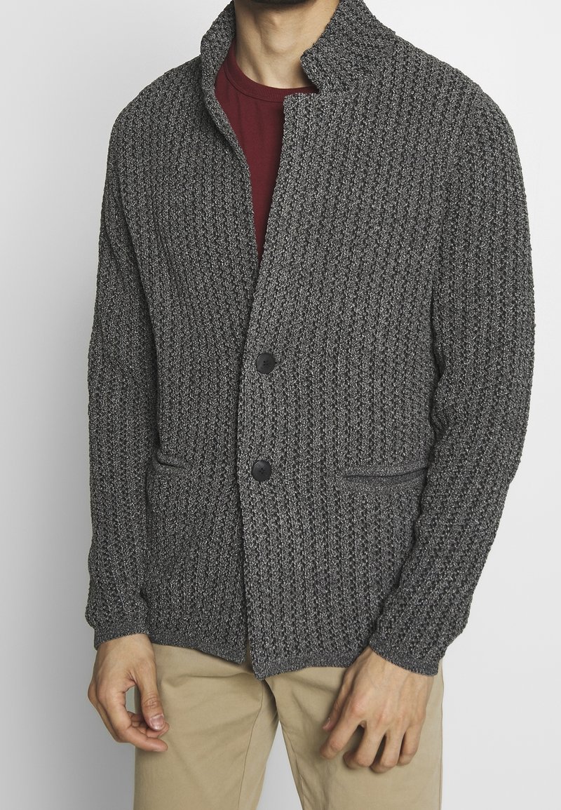 Szara teksturowana dzianinowa kurtka z kołnierzem, dwiema przednimi kieszeniami i zapięciem na guziki. Nosi się ją na bordowej koszuli.