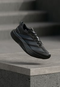 Sapato desportivo preto com design elegante, apresentando uma sola texturada, três riscas escuras e atacadores pretos. O material parece sintético.