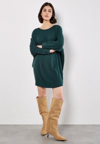 Teal oversized trui-jurk met een gestructureerde afwerking, gecombineerd met beige suède laarzen tot de knieën en een spitse neus. Minimalistisch ontwerp, lange mouwen.