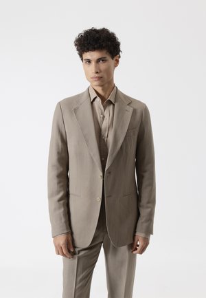 Joven con cabello rizado, vestido con un traje beige y camisa a juego, de pie contra un fondo blanco liso, mirando hacia adelante.