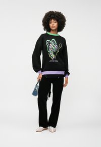 Sweatshirt preto com um gráfico colorido de borboleta e o texto "SANTA CRUZ", apresentando um colarinho verde e uma bainha em lilás. Usado com calças pretas e uma bolsa azul.