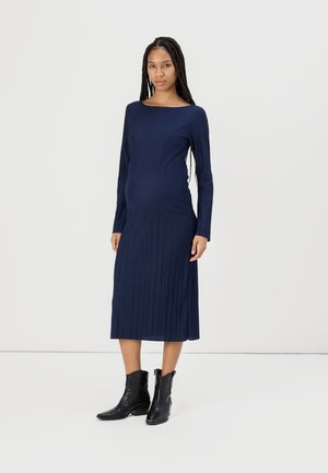 Robe midi côtelée marine avec des manches longues et un col bateau, silhouette ajustée, fentes latérales, associée à des bottines noires.