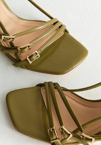 Sandales à talons hauts en cuir verni vert avec un bout carré, un design à plusieurs brides avec des accents de boucles en or, et une semelle intérieure lisse beige.